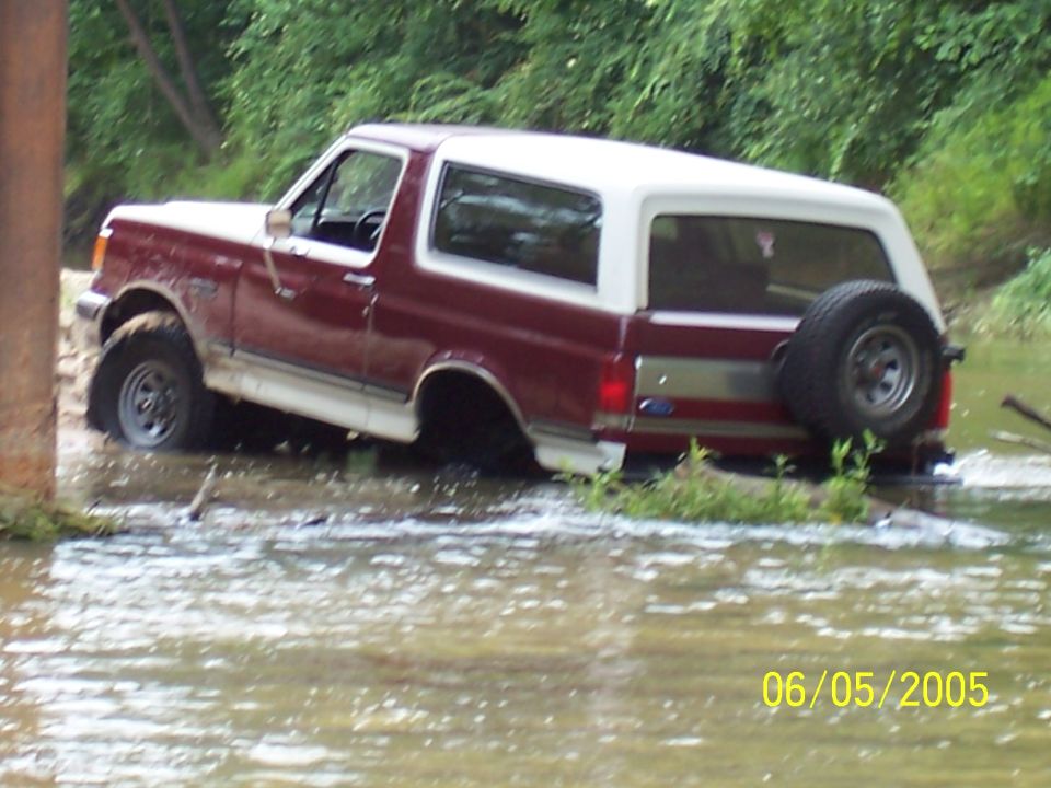 Maroon and white 1990 Ford Bronco partially submerged in water, driving off-road