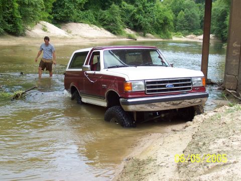 1990 Ford Bronco - A two-tone Ford Bronco navigates a muddy riverbed under a bridge, with a person wading nearby.