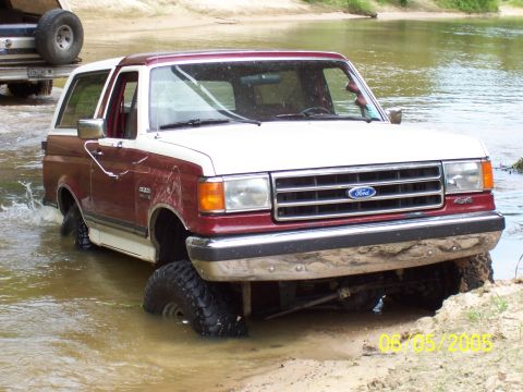 1990 Ford Bronco - A two-tone Ford Bronco navigates a shallow water crossing with its front-left tire deep in the water.
