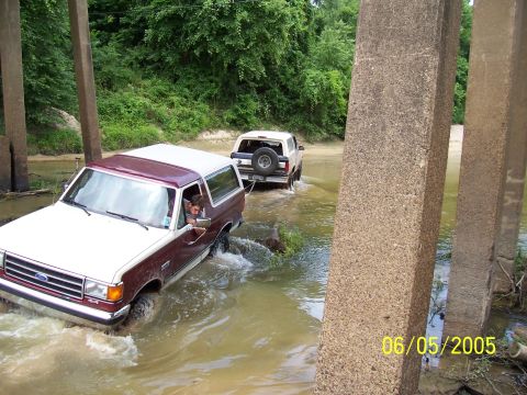 1990 Ford Bronco - Two Ford Broncos navigate a shallow water crossing beneath a bridge.