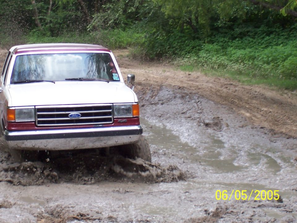 White and maroon 1990 Ford Bronco front view driving through mud