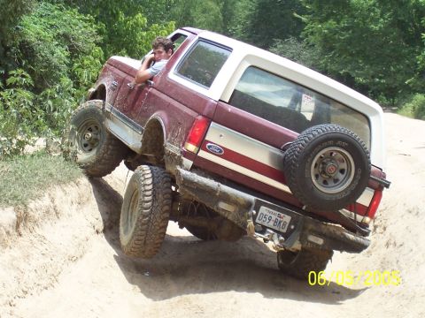 1990 Ford Bronco - A man drives a Ford Bronco off-road, navigating a steep incline with one tire lifted.