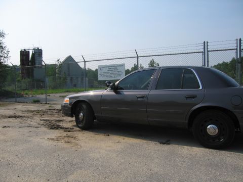 2001 Ford Crown Victoria - A gray 2001 Ford Crown Victoria is parked near a chain-link fence with a sign and industrial buildings in the background.