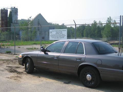 2001 Ford Crown Victoria - A gray 2001 Ford Crown Victoria police car is parked in front of a fenced industrial site with a sign about a Superfund site.