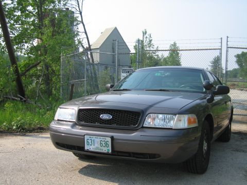 2001 Ford Crown Victoria - Front three-quarter view of a grey unmarked Ford Crown Victoria Police Interceptor parked on gravel next to a chain-link fence with a "Haza…