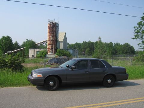 2001 Ford Crown Victoria - Side profile of a charcoal-gray Ford Crown Victoria with black steel police-style wheels and an A-pillar spotlight, parked along a paved ro…
