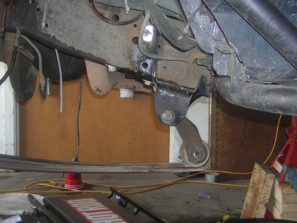 Underside of vehicle showing rusty frame, leaf spring and shackle mount in a garage