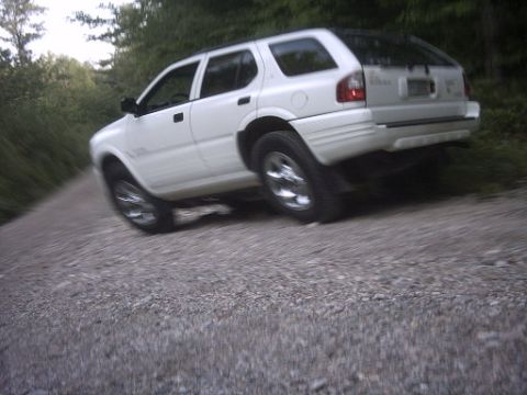 2000 Isuzu Rodeo - White 2000 Isuzu Rodeo SUV driving on a gravel road with trees in the background.
