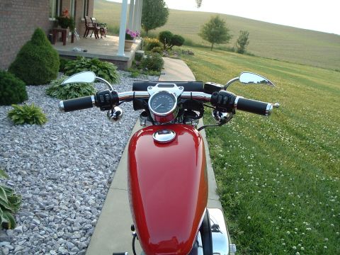 2002 Harley-Davidson Sportster Custom - A red motorcycle is parked on a gravel driveway in front of a house with a grassy hill in the background.