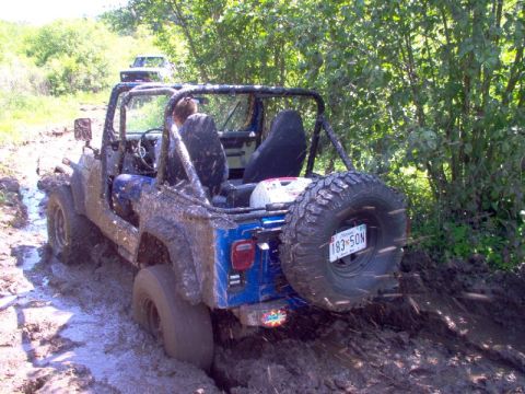 1976 GMC C/K Pickup - A blue Jeep Wrangler is stuck in thick mud on an off-road trail, with another vehicle visible in the background.