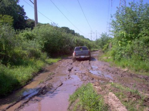 1976 GMC C/K Pickup - A pickup truck navigates a challenging muddy path surrounded by overgrown vegetation and power lines.
