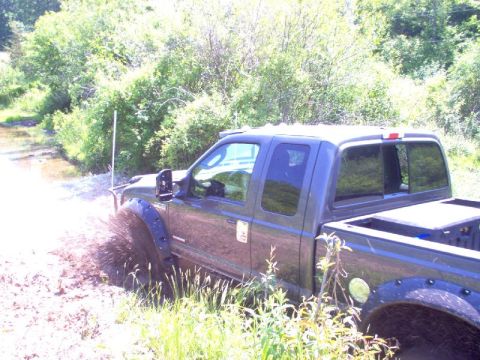1976 GMC C/K Pickup - A dark grey pickup truck navigates a muddy path, kicking up water and dirt.