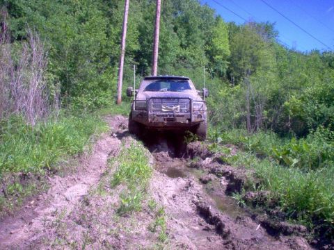 1976 GMC C/K Pickup - A truck navigates a challenging muddy path surrounded by lush green foliage and utility poles.