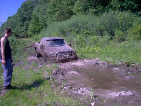 1976 GMC C/K Pickup - A man watches a mud-covered pickup truck stuck in a muddy ditch on a dirt road.