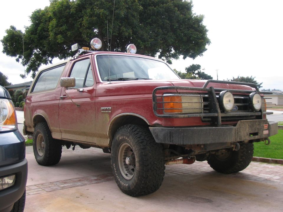 Red lifted Ford Bronco with brush guard and off-road lights, front 3/4 view in driveway