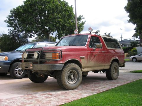 1989 Ford Bronco - This red 1989 Ford Bronco, equipped with a brush guard and auxiliary lights, is parked on a brick driveway next to another vehicle.