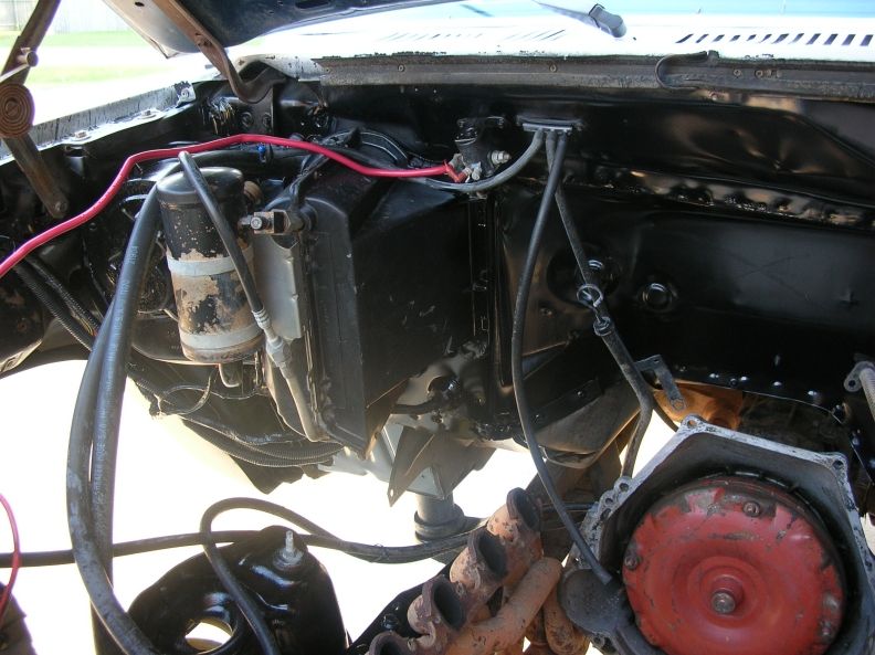 Engine bay of a Ford Bronco showing black firewall, heater box, wiring, distributor, and rusty exhaust manifold