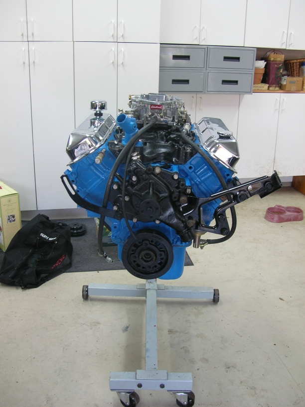 Blue V8 engine with chrome valve covers and Edelbrock carb on an engine stand in a garage
