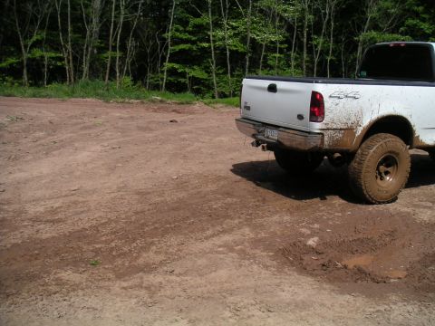 1994 Ford Bronco - A muddy white pickup truck sits on a dirt road surrounded by trees.