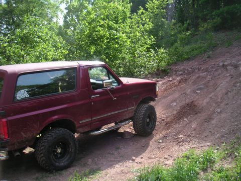 1994 Ford Bronco - A maroon Ford Bronco navigates a steep, rocky dirt path surrounded by trees.