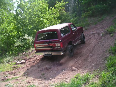1994 Ford Bronco - A red 1994 Ford Bronco navigates a steep, dusty off-road trail surrounded by trees.