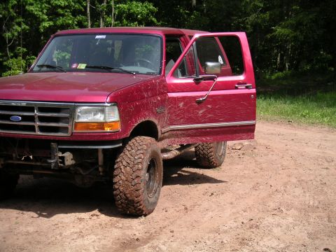 1994 Ford Bronco - A muddy red Ford Bronco sits on a dirt road surrounded by trees, with the driver's door ajar.