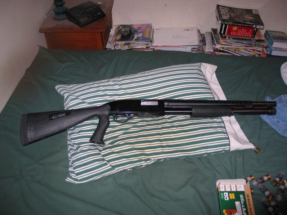 Black pump-action shotgun lying on a striped pillow on a green comforter. Box of shotgun shells visible in the lower right