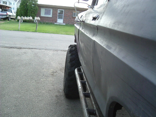Side view of primer-gray lifted pickup on large mud tire with tube side steps, parked on residential street
