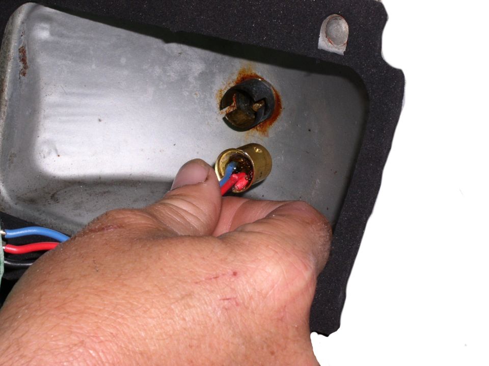 Hand with red and blue wires connecting to a brass bulb socket inside a rusty metal housing with black foam trim