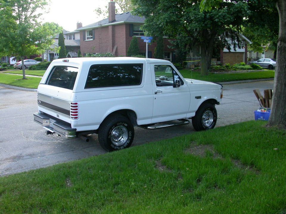 White Ford Bronco SUV parked in a driveway, rear three-quarter view with chrome side steps and white-letter tires