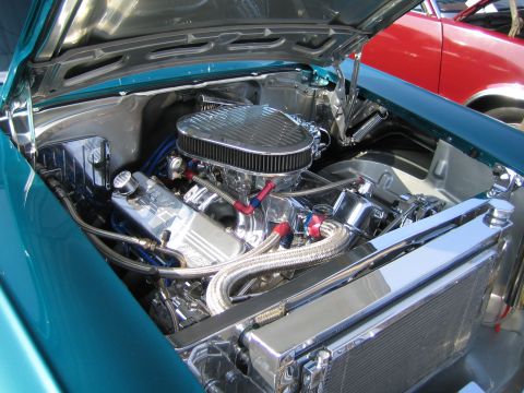 1985 Ford Mustang - The engine bay of a custom car features a polished V8 engine with chrome accents and braided hoses, with another vehicle in the background.