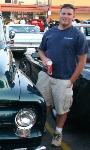 1990 Ford Bronco - young man in a blue shirt and tan cargo shorts holds a drink while leaning against a dark green classic car at an evening car show.