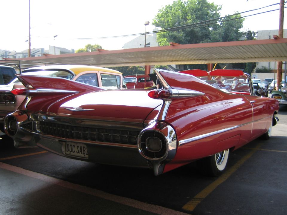 Red convertible with prominent tail fins and chrome bumper. License plate reads 'DOC SAB'. Another car with a yellow roof is behind it