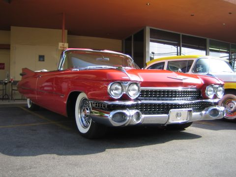 1985 Ford Mustang - A red 1959 Cadillac convertible is parked next to another vehicle under an overhang.
