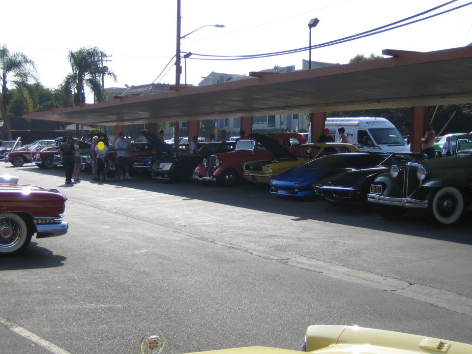 Several classic cars are parked in a row under a covered area. Some have hoods open. People are standing around them