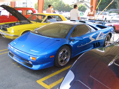 1985 Ford Mustang - A blue sports car is parked in a lot next to other vehicles, including a yellow classic car with its hood open.