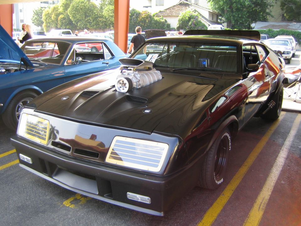 Black custom car with exposed supercharger, wide tires, and spoiler. Another blue car is visible to the left