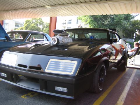 1985 Ford Mustang - A black coupe with a large supercharger protruding from the hood is parked next to another vehicle.