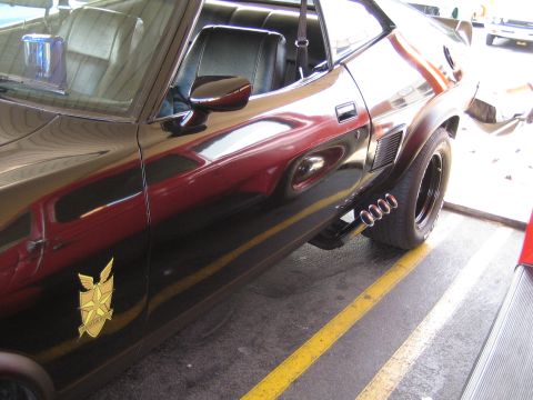 1985 Ford Mustang - Side view of a black coupe with custom exhaust pipes and a gold emblem on the fender.