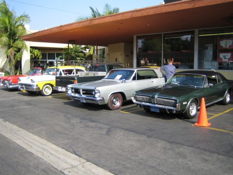 1985 Ford Mustang - A line of classic cars, including a silver Pontiac and a green Mercury Cougar, are parked under an overhang.