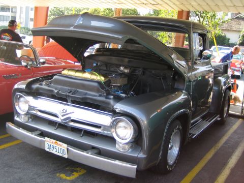 1985 Ford Mustang - A dark gray 1956 Ford F-100 pickup truck is displayed with its hood open, revealing the engine. Another classic car is parked nearby.