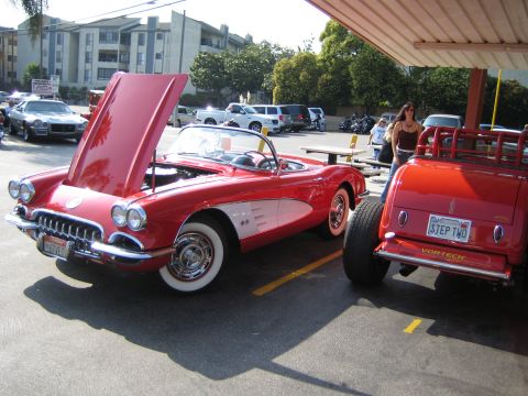 1985 Ford Mustang - A red 1958 Chevrolet Corvette convertible is parked with its hood open, next to another red custom car.