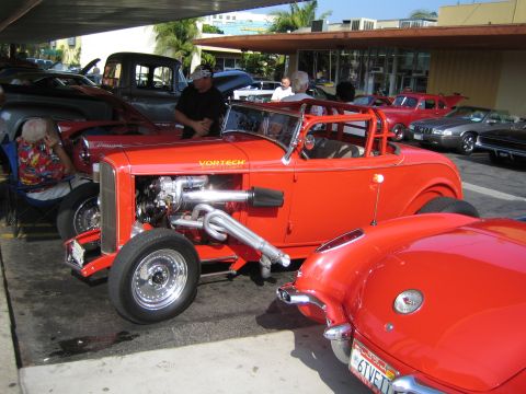 1985 Ford Mustang - A custom red hot rod with a prominent engine and exhaust system is displayed alongside other vehicles.