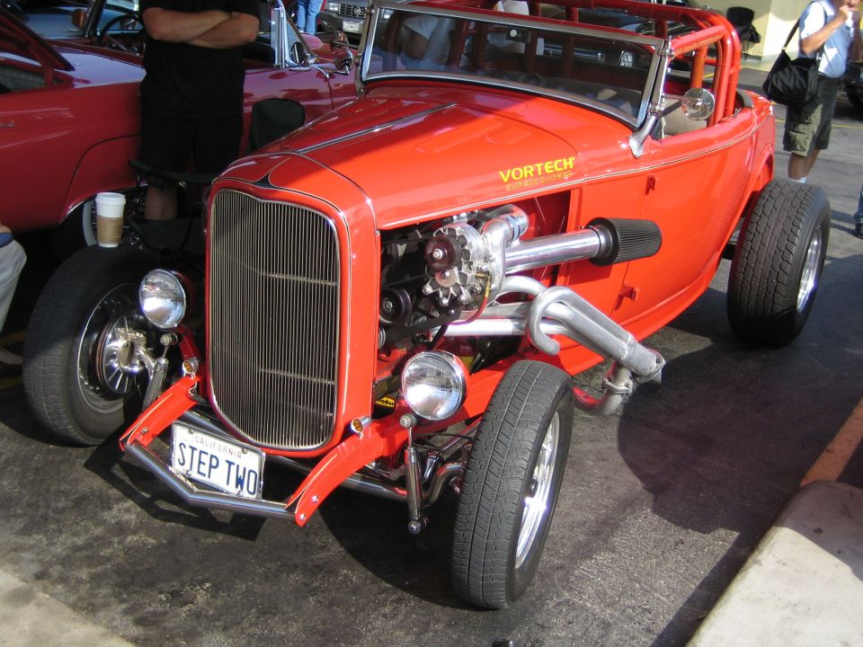 Red hot rod with exposed engine, roll cage, and custom exhaust. License plate reads 'STEP TWO'