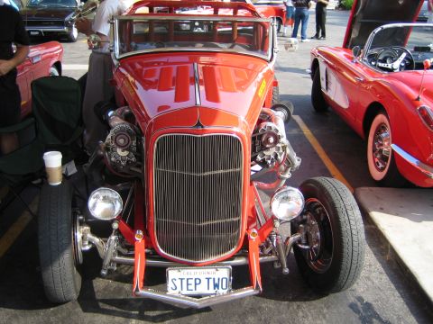 1985 Ford Mustang - A custom red hot rod with prominent dual superchargers is parked next to a red Chevrolet Corvette.