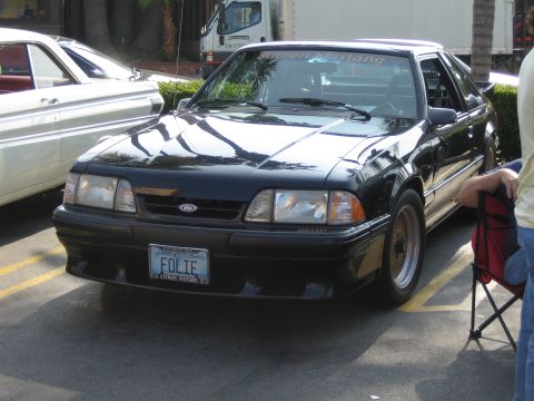 1985 Ford Mustang - A black Ford Mustang is parked in a lot, with another car to its left and a person in a chair to its right.