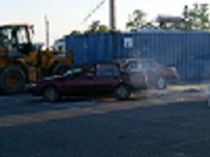 1986 Ford F-250 - A red sedan is seen in a junkyard setting, with heavy machinery and a shipping container nearby.
