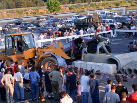 1986 Ford F-250 - A red SUV is being lifted by a front loader as part of an event, with spectators watching.