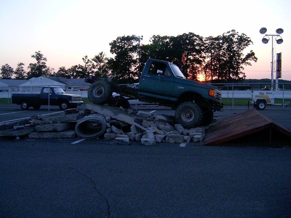 Green pickup truck attempting an obstacle course, with a black pickup truck parked nearby. Sunset in the background