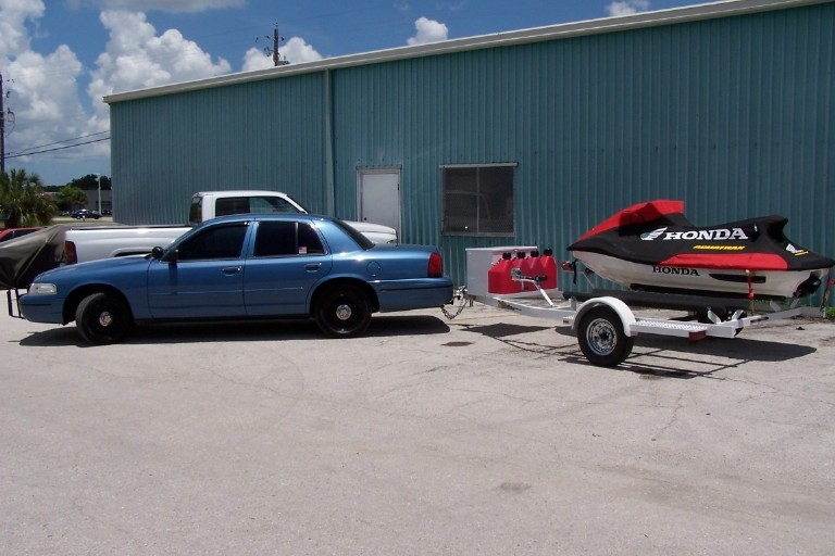 Red and black Honda AquaTrax jet ski on trailer behind a blue sedan, parked by a teal building.